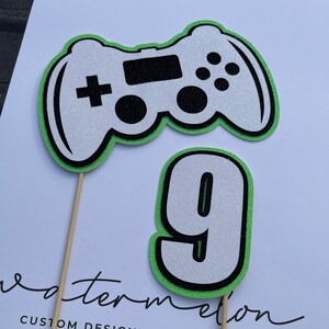 May include: Green and white glitter game controller and a white glitter number nine on wooden sticks. The sticks are on a white surface with the text "watermelon custom designed accessories www.watermelonbyclaire.etsy.com".