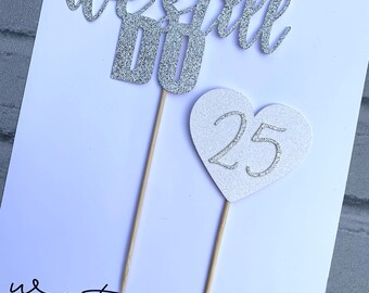 May include: Silver glitter cake topper set with the words "We Still Do" and a heart with the number "25" inside. The topper is on a white background with the text "watermelon custom designed accessories".