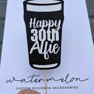 K&ouml;nnte beinhalten: Schwarzer Glitzer-Kuchentopper in Form eines Bierglases mit dem Text "Happy 30th Alfie" in wei&szlig;em Glitzer. Der Topper ist auf einem Holzstiel.
