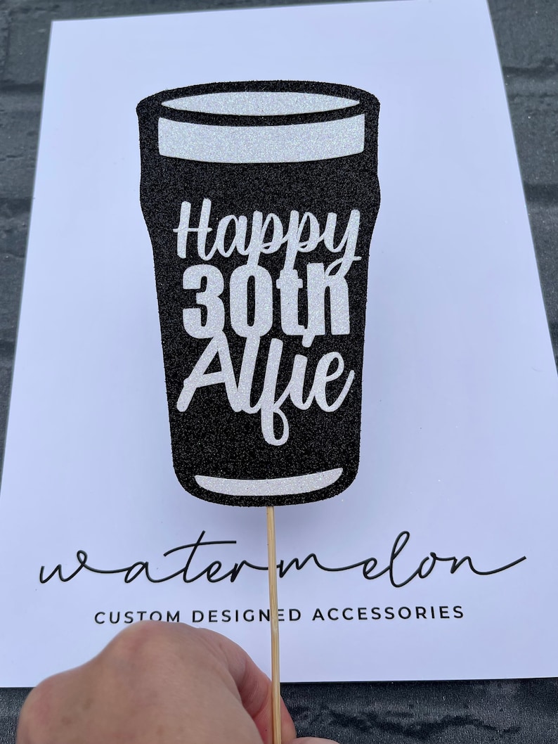 K&ouml;nnte beinhalten: Schwarzer Glitzer-Kuchentopper in Form eines Bierglases mit dem Text "Happy 30th Alfie" in wei&szlig;em Glitzer. Der Topper ist auf einem Holzstiel.