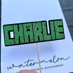 Peut inclure: Topper de gâteau vert pailleté avec le nom "CHARLIE" dans une police pixelisée, sur un fond blanc avec le texte "accessoires conçus par watermelon".