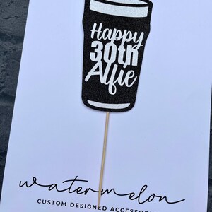 K&ouml;nnte beinhalten: Schwarzer Glitzer-Kuchentopper in Form eines Bierglases mit dem Text "Happy 30th Alfie" in wei&szlig;em Glitzer. Der Topper ist auf einem Holzstiel.