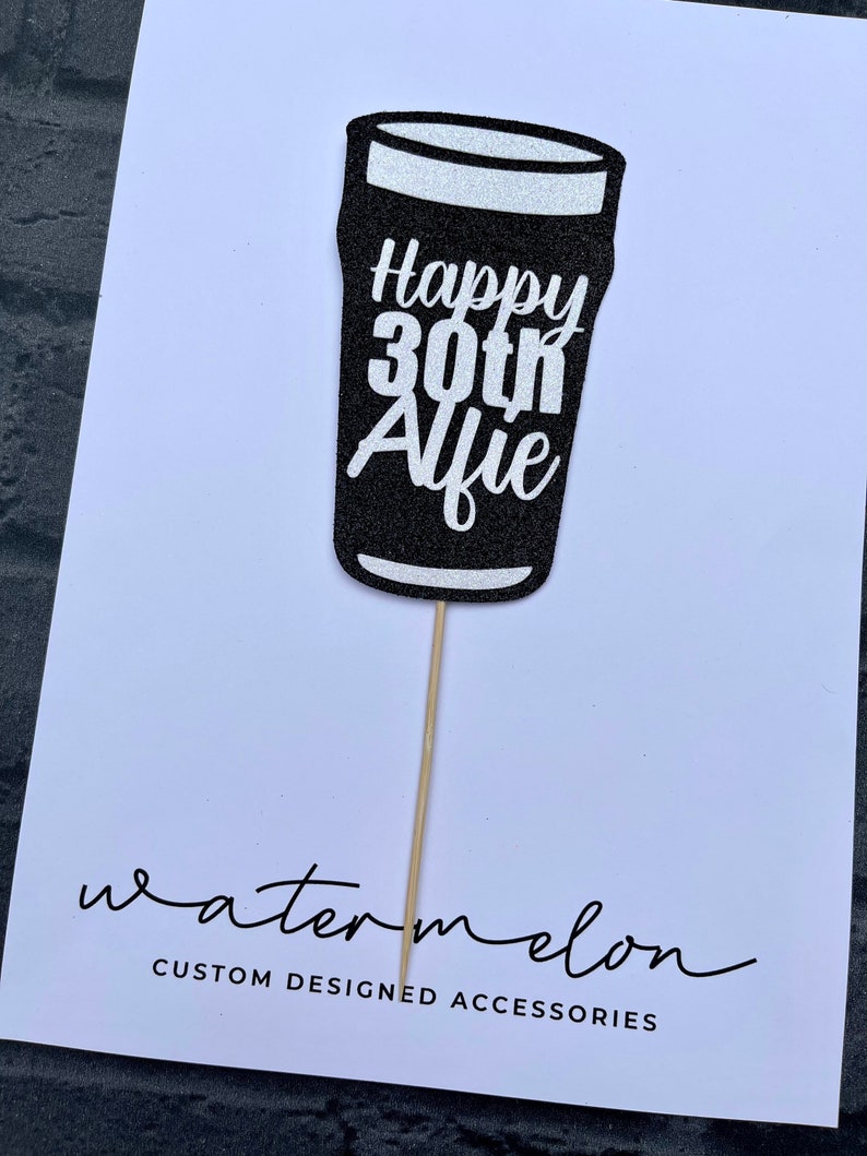 K&ouml;nnte beinhalten: Schwarzer Glitzer-Kuchentopper in Form eines Bierglases mit dem Text "Happy 30th Alfie" in wei&szlig;em Glitzer. Der Topper ist auf einem Holzstiel.