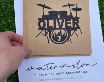Op de afbeelding: Een zwarte en bruine papieren taarttopper met een drumstel silhouet. Het drumstel is voorzien van de naam "OLIVER" en het nummer "7". De tekst "watermelon CUSTOM DESIGNED ACCESSORIES www.watermelonbyclaire.etsy.com" is onder de taarttopper afgedrukt.
