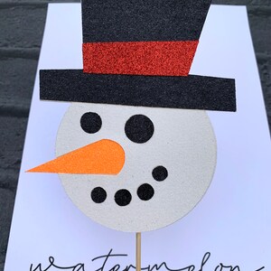 May include: A white snowman face with black eyes and a black mouth. The snowman has an orange carrot nose and is wearing a black top hat with a red band. The snowman is on a wooden stick.