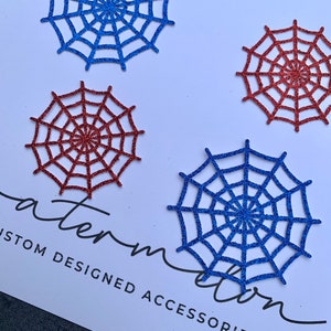 May include: Four glitter spiderwebs, two blue and two red, on a white background. The webs are cut out and have a sparkly finish. The text "Custom Designed Accessories" is printed in black at the bottom of the image.