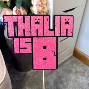 Peut inclure: Un topper de gâteau rose pailleté sur le thème de Minecraft avec le nom "Thalia is 8" en lettres noires.