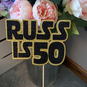 May include: Black and gold glitter cake topper with the text "Russ is 50".