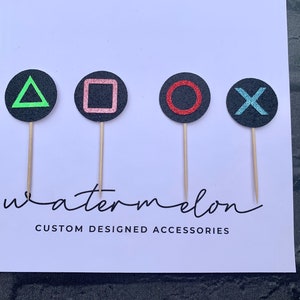 May include: Four black circle cake toppers with different coloured symbols: a green triangle, a pink square, a red circle, and a blue X. The toppers are on wooden sticks and have the text "watermelon CUSTOM DESIGNED ACCESSORIES" below them.