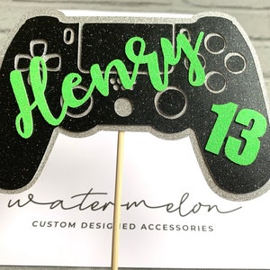 Könnte beinhalten: Schwarzer und silberner Glitzer-Gamecontroller-Kuchentopper mit grüner Beschriftung "Henry 13".