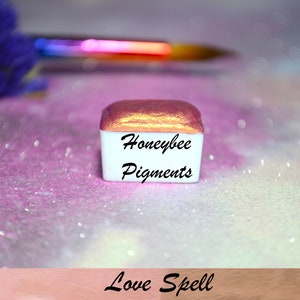 Może przedstawiać: Mały, kwadratowy pojemnik z pigmentem metalicznym w kolorze różowego złota z etykietą "Honeybee Pigments Love Spell".