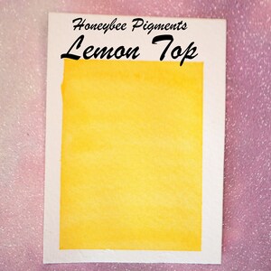 May include: A watercolour swatch of "Lemon Top" pigment from Honeybee Pigments. The swatch is a vibrant, solid yellow colour, set against a white border. The text "Lemon Top" is written in a bold, black script.