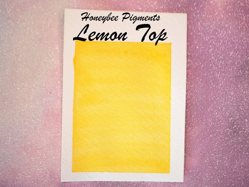 May include: A watercolour swatch of "Lemon Top" pigment from Honeybee Pigments. The swatch is a vibrant, solid yellow colour, set against a white border. The text "Lemon Top" is written in a bold, black script.