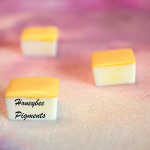 May include: Three small, square containers with a white base and a yellow lid are shown against a soft, pink background. The text "Honeybee Pigments" is printed on one of the containers. The items appear to be art supplies.