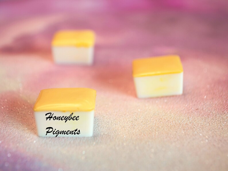 May include: Three small, square containers with a white base and a yellow lid are shown against a soft, pink background. The text "Honeybee Pigments" is printed on one of the containers. The items appear to be art supplies.