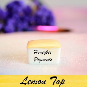 May include: A small, square container of "Lemon Top" pigments from Honeybee Pigments. The container has a white base and a light yellow lid. The background is a soft pink and white with purple flowers and a paintbrush.