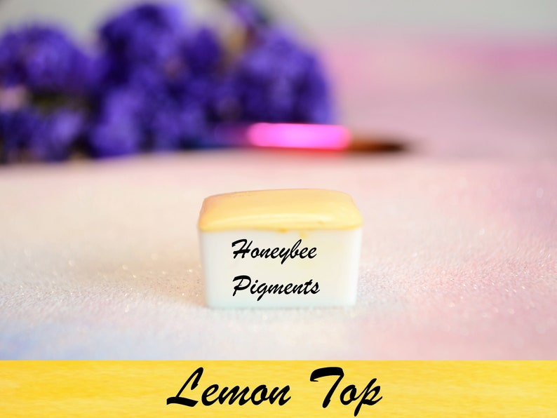 May include: A small, square container of "Lemon Top" pigments from Honeybee Pigments. The container has a white base and a light yellow lid. The background is a soft pink and white with purple flowers and a paintbrush.