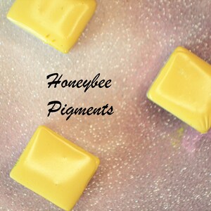 May include: Three yellow, diamond-shaped pigments are scattered on a sparkly, pink surface. The text "Honeybee Pigments" is written in black cursive. The pigments have a smooth, glossy finish.