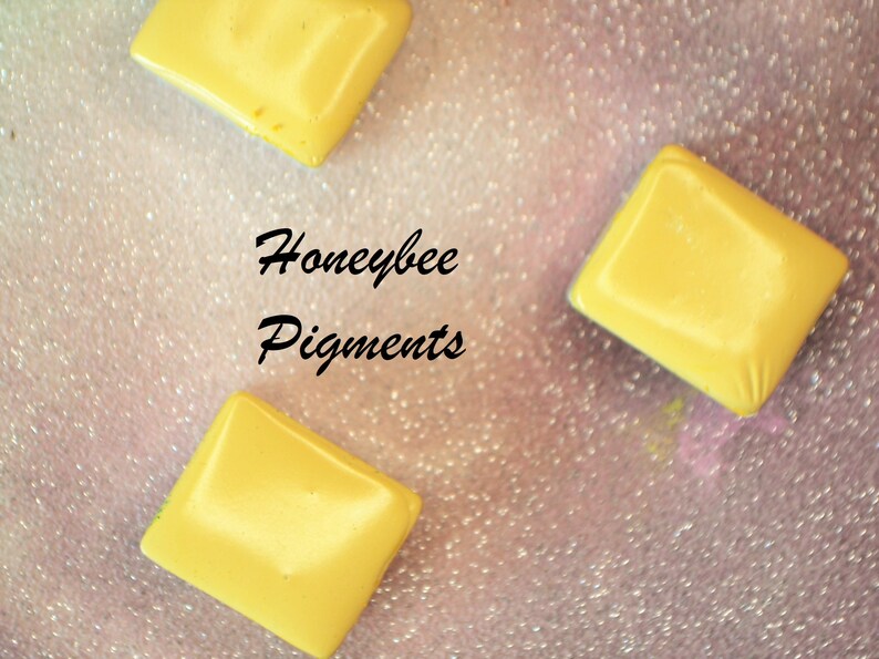 May include: Three yellow, diamond-shaped pigments are scattered on a sparkly, pink surface. The text "Honeybee Pigments" is written in black cursive. The pigments have a smooth, glossy finish.