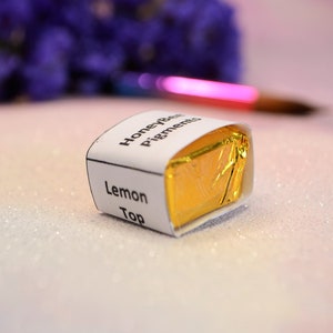 May include: A small, square package of HoneyBee Pigments Lemon Top, wrapped in gold foil and a white label. The label displays the product name. A paintbrush and purple flowers are in the background.