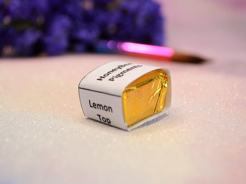 May include: A small, square package of HoneyBee Pigments Lemon Top, wrapped in gold foil and a white label. The label displays the product name. A paintbrush and purple flowers are in the background.