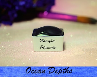Op de afbeelding: Een kleine, vierkante witte container met een zwart deksel. De container is gelabeld "Honeybee Pigments" en de tekst "Ocean Depths" is in blauw onder de container geschreven.