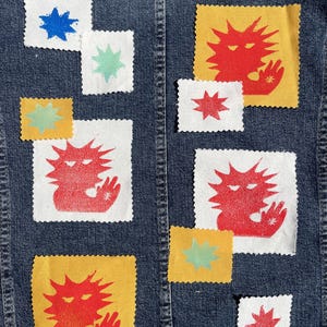 Op de afbeelding: De achterkant van een spijkerjasje met verschillende stoffen patches. De patches hebben rode, blauwe en groene sterren en creatuurontwerpen op witte en gele achtergronden. De jas is een medium blauwe wassing.