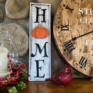 May include: A white wooden sign with the word "HOME" in black letters. The letter "O" is replaced with a carved orange pumpkin. The sign is on a wooden shelf with a red cardinal figurine and a wooden clock.