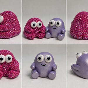 May include: Two small figurines with large googly eyes. One figurine is covered in tiny, shimmering pink beads, while the other is smooth, metallic lavender with a simple black line for a mouth. Both have rounded bodies and small limbs.