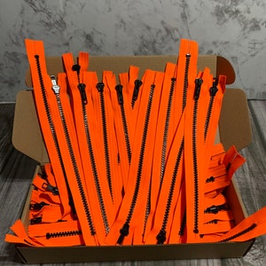 May include: A box filled with bright orange zippers with black pulls. The zippers are arranged in a neat pile.