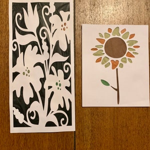 May include: Two paper cutouts of flowers. The first cutout is a white floral design on a black background. The second cutout is a brown, green, and orange sunflower.