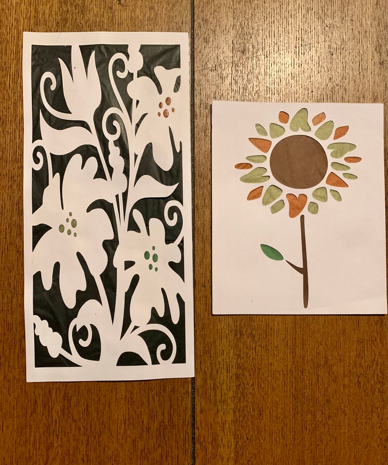 May include: Two paper cutouts of flowers. The first cutout is a white floral design on a black background. The second cutout is a brown, green, and orange sunflower.