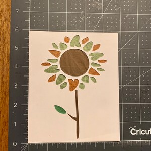 May include: A white vinyl stencil of a sunflower with brown and green petals and a brown stem. The stencil is on a gray cutting mat.