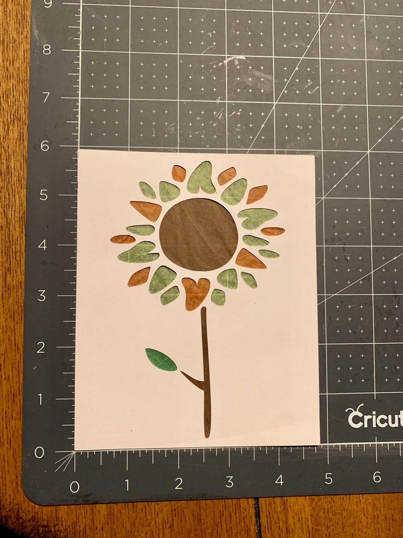 May include: A white vinyl stencil of a sunflower with brown and green petals and a brown stem. The stencil is on a gray cutting mat.