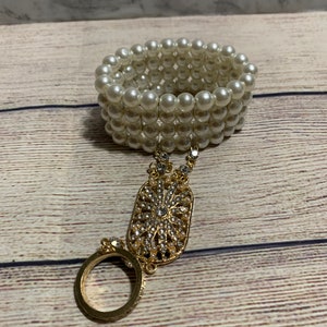 Op de afbeelding: Een parelarmband verbonden met een gouden ring met een decoratief gouden en strass middenstuk. De armband is gemaakt van meerdere strengen witte parels. De ring en het middenstuk zijn goudkleurig.