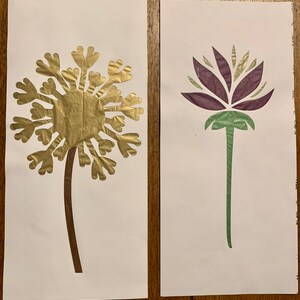 May include: Two paper cutouts of flowers on white paper. The first is a gold dandelion with a brown stem. The second is a purple flower with a green stem.