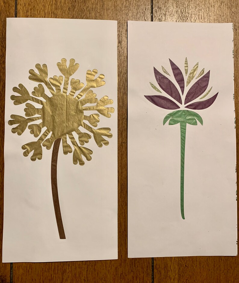 May include: Two paper cutouts of flowers on white paper. The first is a gold dandelion with a brown stem. The second is a purple flower with a green stem.