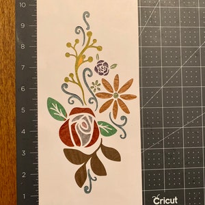 May include: A white stencil with a floral design featuring a red rose, brown leaves, a yellow flower with brown petals, and a purple flower with green leaves.