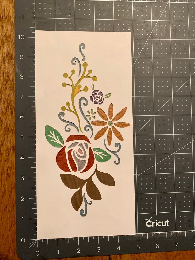 May include: A white stencil with a floral design featuring a red rose, brown leaves, a yellow flower with brown petals, and a purple flower with green leaves.
