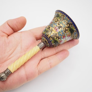 May include: A small, silver bell with a white, twisted handle. The bell is decorated with colourful enamel and floral patterns.