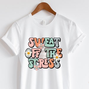 May include: White t-shirt with a retro-style graphic that says "Sweat Off The Stress" in colorful letters with flowers.