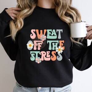 Puede incluir: Sudadera negra con un gr&aacute;fico de estilo retro que dice "Sweat Off The Stress" en letras de colores con flores.