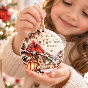 Peut inclure: Une d&eacute;coration de No&euml;l ronde en c&eacute;ramique illustrant deux cardinaux rouges sur une branche de pin enneig&eacute;e avec une lanterne et un ornement en forme de c&oelig;ur. Le texte indique Our First Christmas Together 2026. Suspendue par une ficelle.