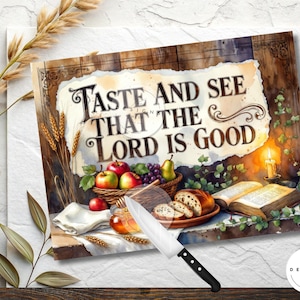 May include: A watercolour print with the words "Taste and See That the Lord is Good." The image shows a basket of fruit, bread, a lit candle, and an open book. A knife is in the foreground. The Designs Nest logo is in the bottom right corner.