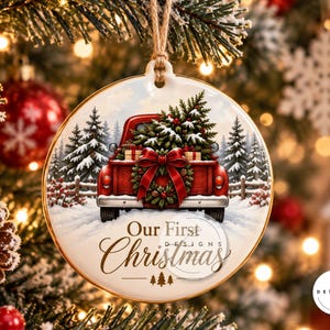 Peut inclure: Une d&eacute;coration de No&euml;l ronde en c&eacute;ramique blanche illustrant un pick-up rouge transportant un sapin enneig&eacute;. Un grand n&oelig;ud rouge orne le hayon. Le texte Our First Christmas est inscrit avec trois petits sapins en dessous. Suspension en corde.