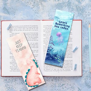 May include: Two watercolor bookmarks with text and illustrations are displayed on an open book. One bookmark says "Just Keep Reading" with a dinosaur. The other says "Learn Something New Today" with a jellyfish.