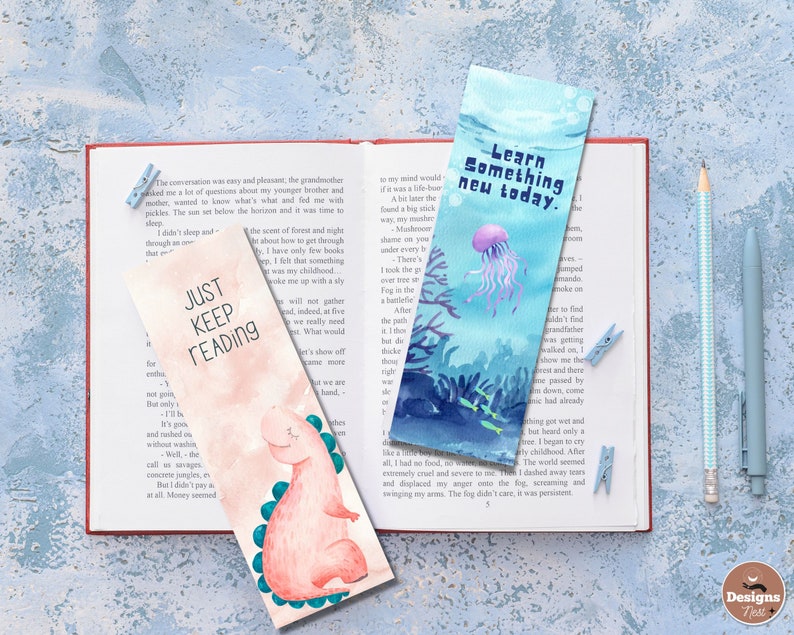 May include: Two watercolor bookmarks with text and illustrations are displayed on an open book. One bookmark says "Just Keep Reading" with a dinosaur. The other says "Learn Something New Today" with a jellyfish.