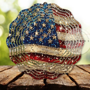 May include: A layered, decorative ornament featuring the American flag design. The ornament has a wavy, layered edge and is decorated with glitter. The flag design includes red and white stripes, blue field with stars, and gold glitter accents. The ornament is displayed on a wooden surface.