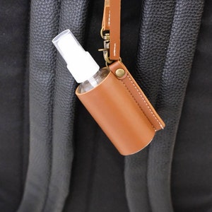 K&ouml;nnte beinhalten: Ein brauner Leder-Handdesinfektionsmittelhalter mit einer wei&szlig;en Spr&uuml;hflasche, die an einem braunen Lederband befestigt ist. Der Halter ist an einem schwarzen Rucksack befestigt.