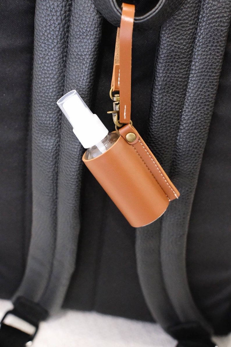 K&ouml;nnte beinhalten: Ein brauner Leder-Handdesinfektionsmittelhalter mit einer wei&szlig;en Spr&uuml;hflasche, die an einem braunen Lederband befestigt ist. Der Halter ist an einem schwarzen Rucksack befestigt.